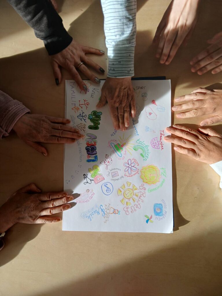 Mehrere Hände liegen auf einem Tisch um ein Blatt mit bunten Zeichnungen und Schriftzügen