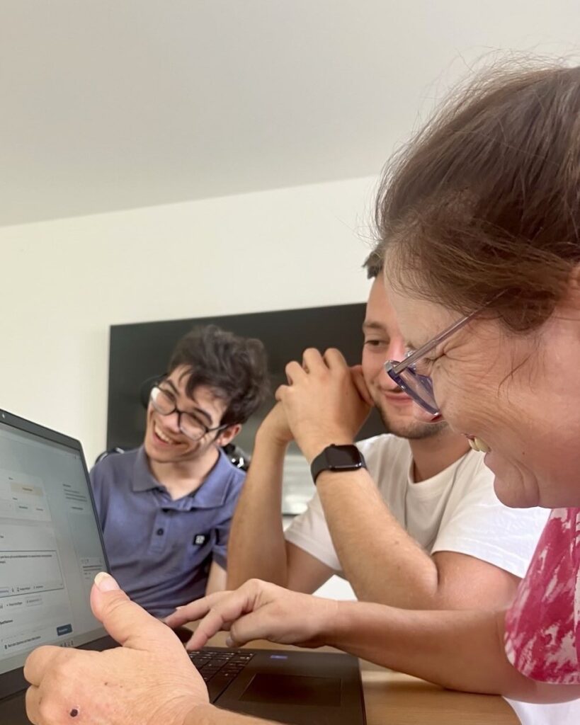 Drei Personen sitzen an einem Tisch und blicken gemeinsam auf einen aufgeklappten Laptop. Eine Person im Vordergrund zeigt mit dem Finger auf den Bildschirm. Die anderen beiden Personen sitzen nebeneinander, eine davon stützt den Kopf auf die Hand und trägt eine helle Armbanduhr. Im Hintergrund ist eine dunkle, rechteckige Fläche an der Wand zu sehen.