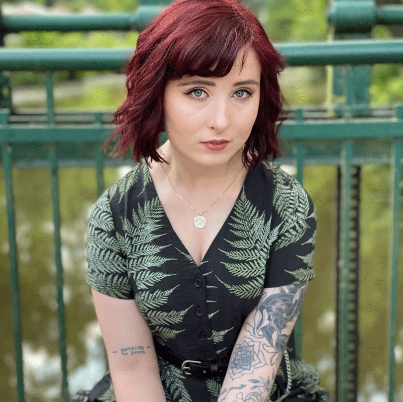 Person mit schulterlangem, rotbraunem Haar trägt ein kurzärmeliges Kleid mit Farnmuster und zeigt einen tätowierten rechten Unterarm sowie ein Armband am linken Handgelenk